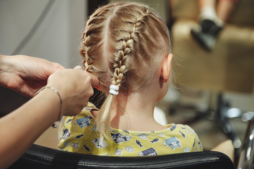 Barber woman make fashionable pretty hairstyle for cute little blond girl child in modern barbershop, hair salon. Hairdresser makes hairdo for young baby in barber shop. Concept hairstyle and beauty