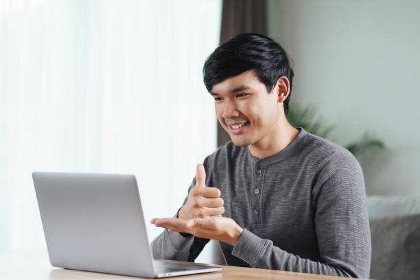 молодой азиатский мужчина глухой инвалид использует ноутбук для онлайн-видеоконференций, обучаясь и общаясь на языке жестов. - sign language стоковые фото и изображения