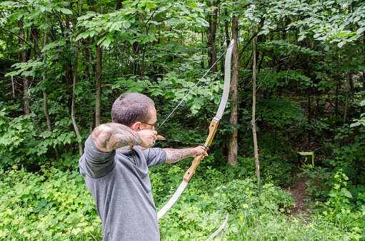 Tattooed young man using a bow and aiming a target in the forest