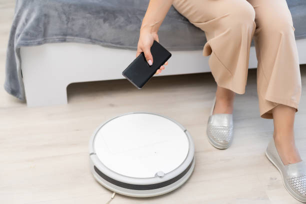 istock Young woman using automatic vacuum cleaner to clean the floor, controling machine housework robot 1332512639