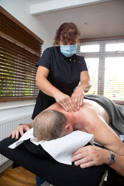 마사지 테이블에 엎드린 남자에게 깊은 조직 마사지를 주는 보호 얼굴 마스크를 착용한 여성 안마사 - massaging deep human tissue pain 뉴스 사진 이미지