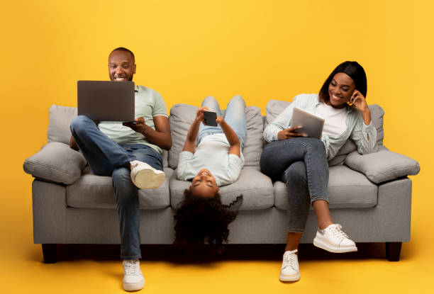 gadgets sucht. junge schwarze dreierfamilie mit verschiedenen elektronischen geräten, die auf dem sofa auf gelbem hintergrund sitzt - family african ethnicity black african descent stock-fotos und bilder