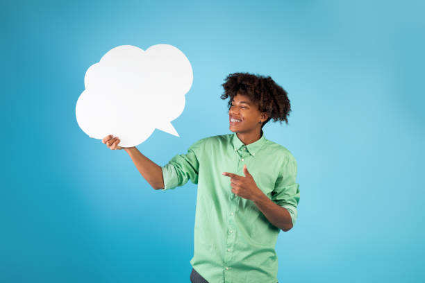 istock Cool african american guy pointing at empty speech bubble and smiling, standing over blue studio background 1332517662