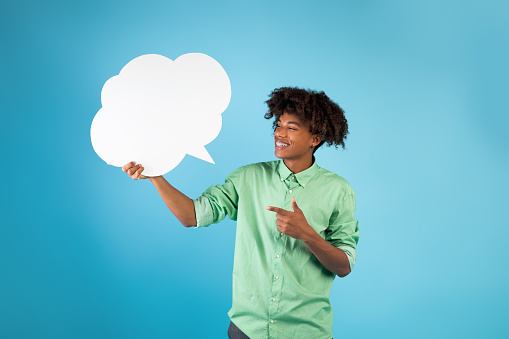 Cool african american guy pointing at empty speech bubble and smiling, standing over blue studio background, mockup for design. Happy teenager with blank word cloud, copy space