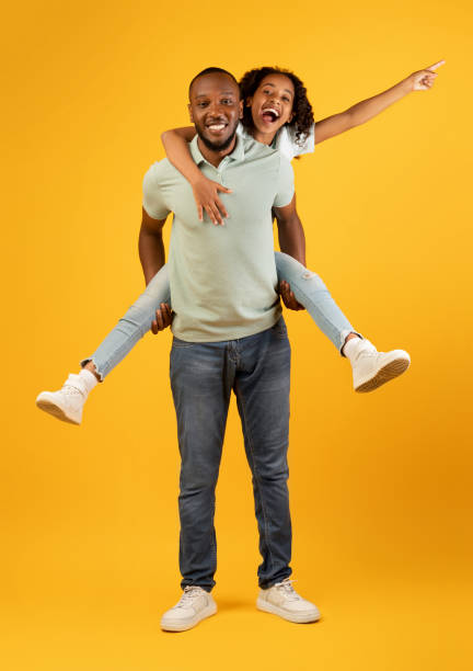 istock Father's love. Exited black dad carrying his daughter on back, having fun and spending time together on yellow background 1332517736