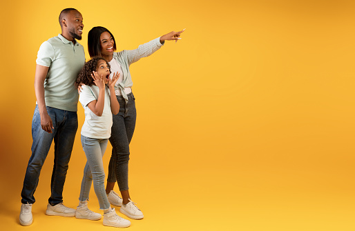 Wow offer. Full body length portrait of excited black family looking and pointing aside at copy space on yellow background, demonstrating free place for your advert, panorama, banner