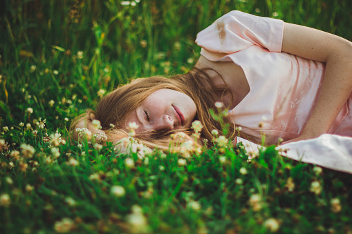 Portrait of a girl with long hair in a pink dress lies and sleeps in the green grass with white clover flowers on a warm summer evening. Photo of beautiful sleeping woman in the field with wildflowers