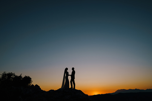 Silhouettes of the bride and groom stand on the mountain on the sunset. High quality photo
