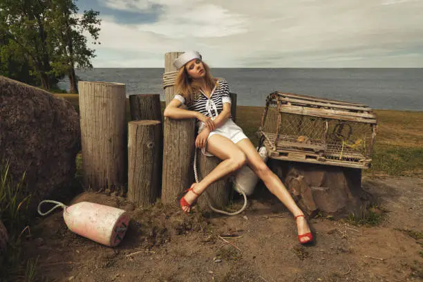 Photo of Young blonde woman with crimped hair, dressed in a sailor style outfit, sitting on wooden pillars in a fisherman's cove