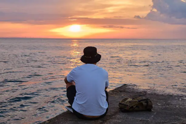 Photo of A young man sits by the sea and looks at the dawn. Male on travels. Amazing picturesque outdoor view. Beautiful dawn on the seashore. Healthy Lifestyle, Leisure Activities. Travel and Exploration