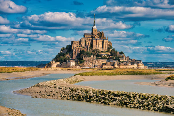 Mont-Saint-Michel Abbey / France Normandy, France, August 29, 2019: Looking at the magestic Mont Saint-Michel. It is seen as a example building of Romanesque architecture. 
Le Mont-Saint-Michel or in english Saint Michael's Mount is a tidal island and mainland commune in Normandy, France. The island lies approximately one kilometre (0.6 miles) off the country's north-western coast, at the mouth of the Couesnon River near Avranches and is 7 hectares (17 acres) in area. The mainland part of the commune is 393 hectares (971 acres) in area so that the total surface of the commune is 400 hectares (988 acres). As of 2017, the island has a population of 30. 
As of 2017, the island has a population of 30. Mont-Saint-Michel and its bay are on the UNESCO list of World Heritage Sites. It is visited by more than 3 million people each year. Over 60 buildings within the commune are protected in France as monuments historiques. mont saint michel photos stock pictures, royalty-free photos & images