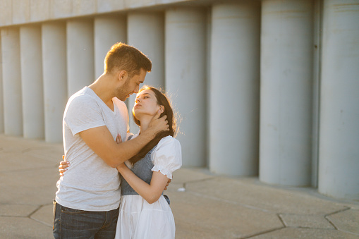 Medium shot portrait of happy romantic young couple in love hugging while man touching neck of girlfriends with hand, in city park at sunset on background of warm bright sunlight.