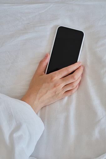 Close-up of a hand holding a smartphone in bed. The girl reaches for her mobile phone to turn off the alarm or read a message. The man fell asleep with a phone in his hand