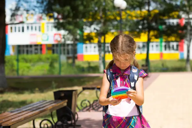 Photo of Little girl with a backpack and in a school uniform in the school yard plays pop it toy. Back to school, September 1. The pupil relaxes after lessons. Primary education, elementary class for student