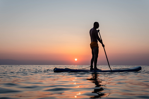 Silhouette of active man standing with paddle on SUP at sunset background. Paddleboard on the beach at sunset