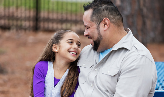 An Hispanic girl at the park with her father. They are sitting side by side, smiling at each other. The daughter is 9 years old and her proud and loving dad is in his 30s.