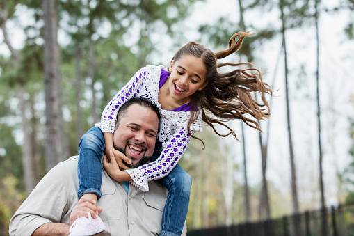 An Hispanic father carrying his daughter on his shoulders at the park. They are playing and laughing, leaning to one side. The girl is 9 years old and dad is in his 30s. She is holding on tight to her father's head so she doesn't fall off.