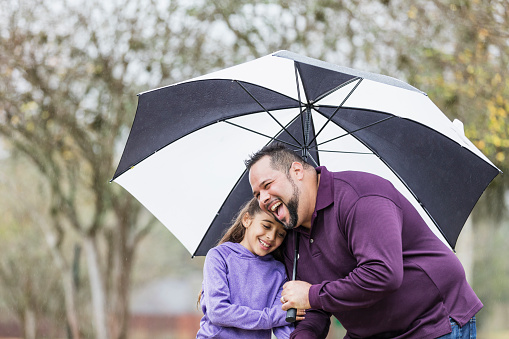 An Hispanic girl and her father in the park on a rainy day, holding hands and laughing. Dad is holding a big umbrella over their heads to keep them dry. The man, in his 30s, is bending down and his 9 year old daughter is resting her head on his shoulder.