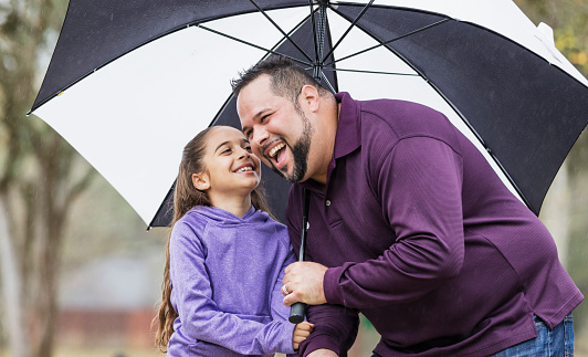 An Hispanic girl and her father in the park on a rainy day, holding hands and smiling. Dad is holding a big umbrella over their heads to keep them dry. The man, in his 30s, is bending down so his 9 year old daughter can tell him something.