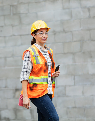 A female construction worker at a job site wearing a hard hat, safety glasses and reflective vest, holding a mobile phone. The Hispanic woman is in her 40s.  She is walking outdoors by a concrete block wall, looking away with a serious expression.