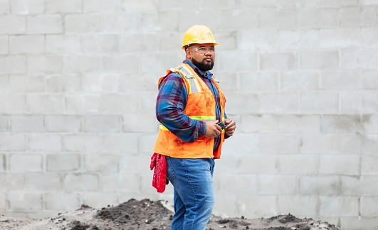 A construction worker at a job site wearing a hard hat, safety glasses and reflective vest, holding a walkie-talkie. He is in his 30s, mixed race African-American and Pacific Islander. He is standing outdoors by a concrete block wall, looking away with a serious expression.