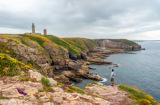 A young tourist taking a photo on the coast next to the Phare Du Cap Frehel, it is a maritime lighthouse in Cotes-d´Armor (France). At the tip of Cap Frehel