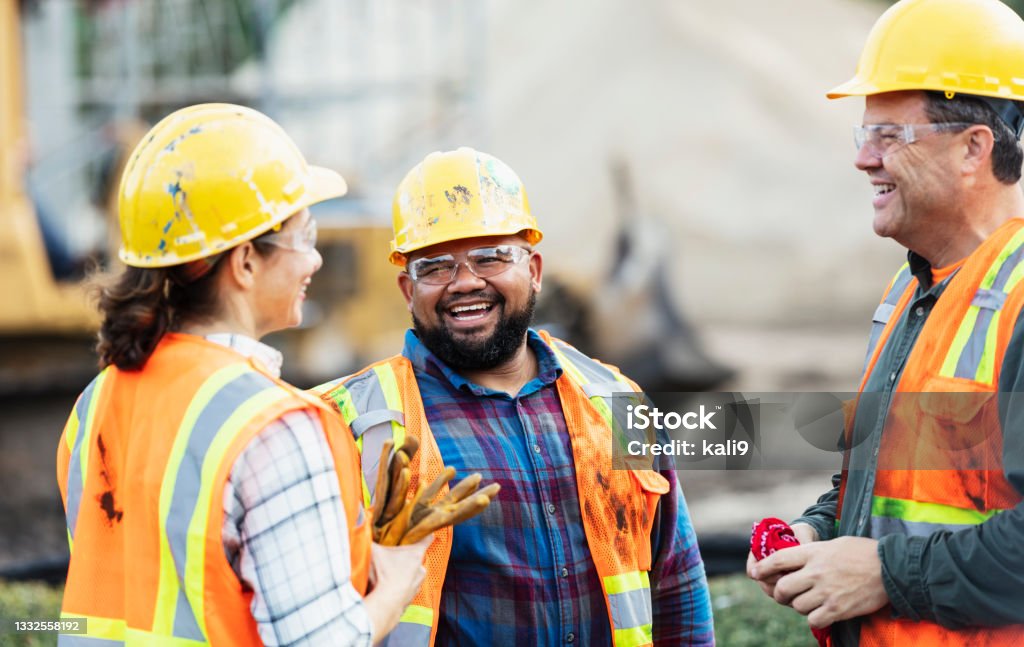 Three multi-ethnic construction workers chatting A group of three multi-ethnic workers at a construction site wearing hard hats, safety glasses and reflective clothing, smiling and conversing. The main focus is on the mixed race African-American and Pacific Islander man in the middle. The other two construction workers, including the woman, are Hispanic. Construction Site Stock Photo