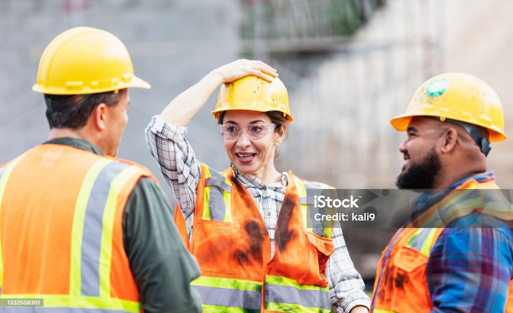 Hispanic woman and two men, workers at construction site A group of three multi-ethnic workers at a construction site wearing hard hats, safety glasses and reflective clothing, smiling and conversing. The Hispanic woman in the middle is smiling, and adjusting her hardhat, looking toward the Hispanic man. Adjusting Stock Photo