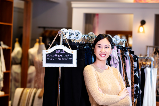 A young Asian woman standing at a clothes rack in a clothing store, next to a SALE sign. She is a saleswoman, or perhaps the owner of this small business. She is smiling confidently at the camera with her arms crossed.