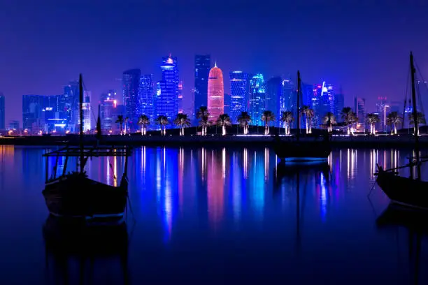 Coloful illuminated skyline of Doha at night with traditional wooden boats called Dhows in the foreground, Qatar, Middle East against dark sky