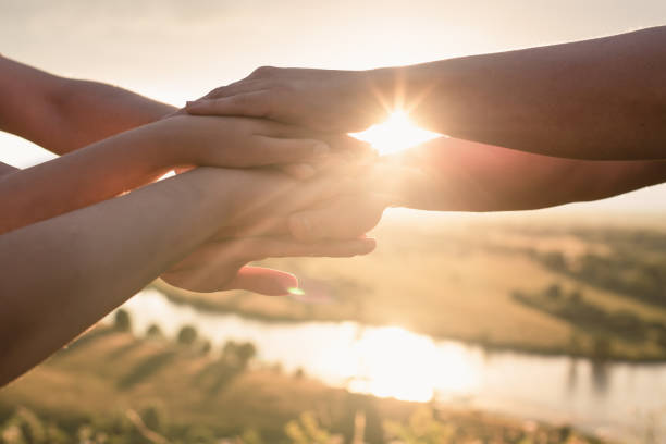 las manos de las personas están dobladas una encima de la otra para desarrollar el equipo y crear un equipo contra el fondo del cielo y el sol poniente. el concepto de unidad, apoyo mutuo y asistencia mutua. - cheering business three people teamwork fotografías e imágenes de stock