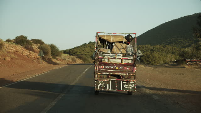 Driving on road behind heavy leaded local small truck in landscape of wild Africa. back view of old car with bizarre tunning. Morocco, Africa