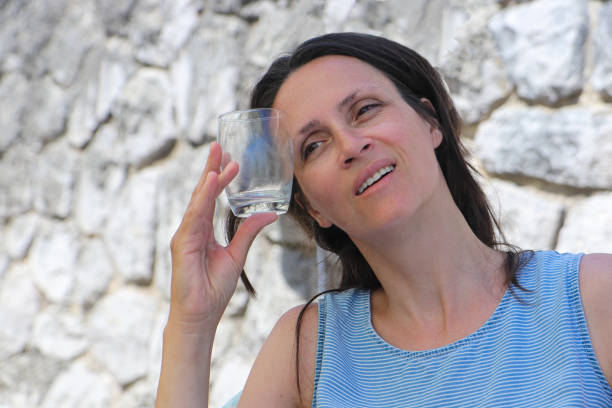 Exhausted woman due to extreme heat cooling down herself by holding cold drinking glass on her forehead.