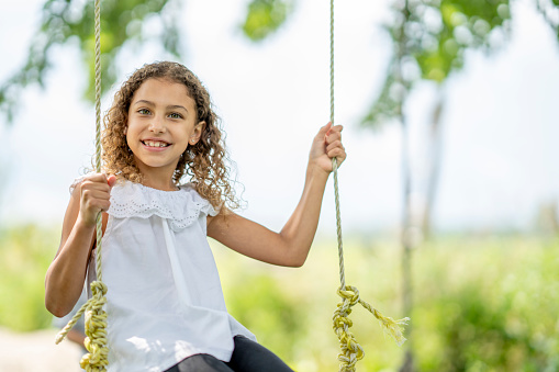 A girl smiles while swinging from a tree in a lovely rural summer setting.