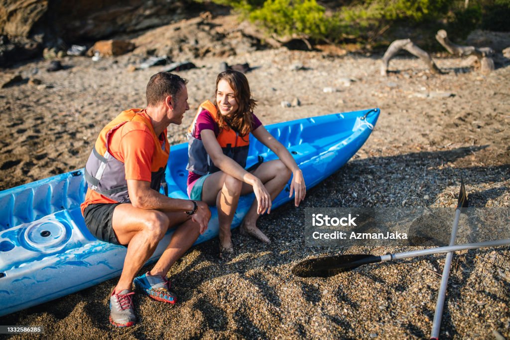 Spanish Kayakers in 30s and 40s After Morning Exercise Male and female friends sitting on side of beached tandem kayak and smiling at each other after morning paddle in Mediterranean Sea off the Costa Brava. Life Jacket Stock Photo