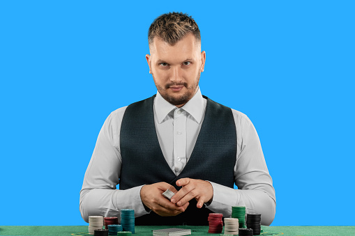 Male croupier at the casino at the table isolated on blue background. Casino concept, gambling, poker, chips on the green casino table.