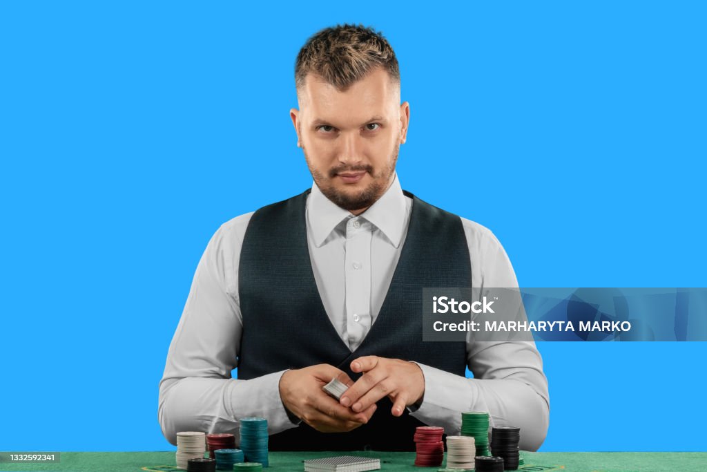 Male croupier at the casino at the table isolated on blue background. Casino concept, gambling, poker, chips on the green casino table. Male croupier at the casino at the table isolated on blue background. Casino concept, gambling, poker, chips on the green casino table Casino Worker Stock Photo