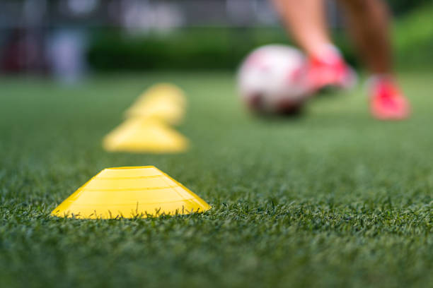 A yellow obstacle cone (Focus), sport training equipment on artificial pitch ground with action of a football player is practice to dribbling the ball as blurred background. Sport training concept.