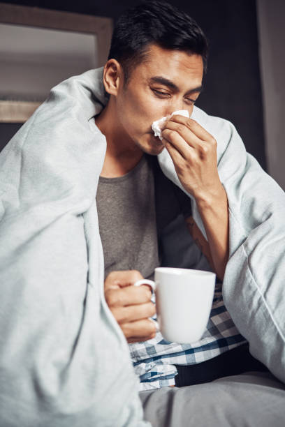 shot of a young man blowing his nose and having tea while recovering from an illness in bed at home - allergy sneezing cold and flu flu virus imagens e fotografias de stock
