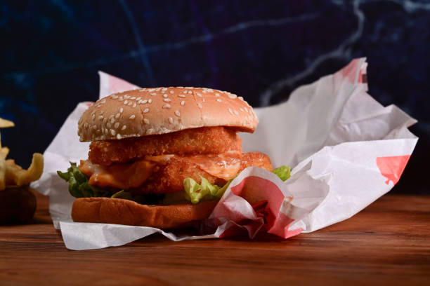istock Delicious fish burger with fresh vegetables and cheese on wooden table. 1332621122