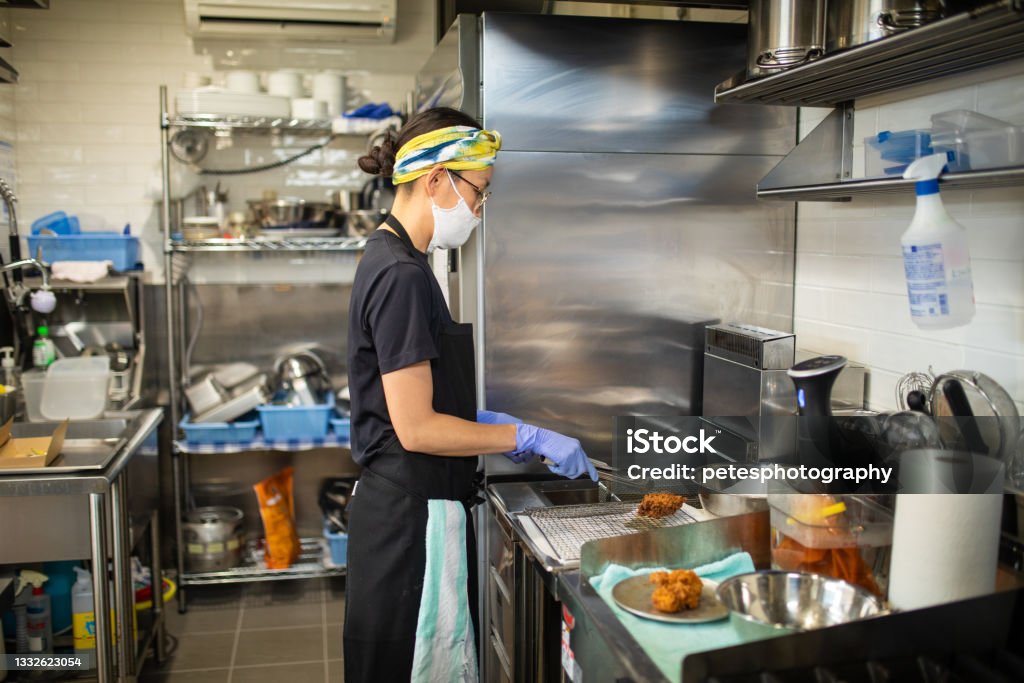 Female chef preparing food in her cafe kitchen A Japanese female chef preparing food in her kitchen wearing protective mask and gloves. Commercial Kitchen Stock Photo
