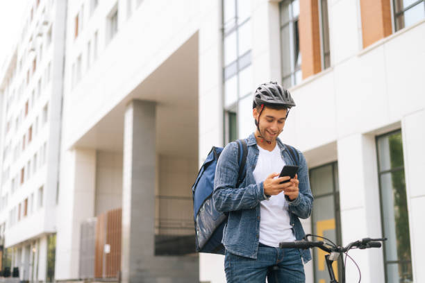 istock Medium shot of cheerful handsome male courier with thermo backpack standing with bicycle in city street and using navigation app on phone. 1332633900