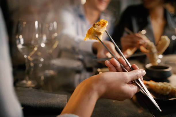 Detail of a woman's hand grabbing a spring roll with metallic chopsticks. He sits at a table with friends in a fancy asiatic fusion food restaurant. stock photo
