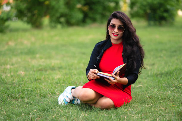 Education, high school and people concept - Outdoor Portrait of young Asian/Indian female student sitting on ground and reading notebook.