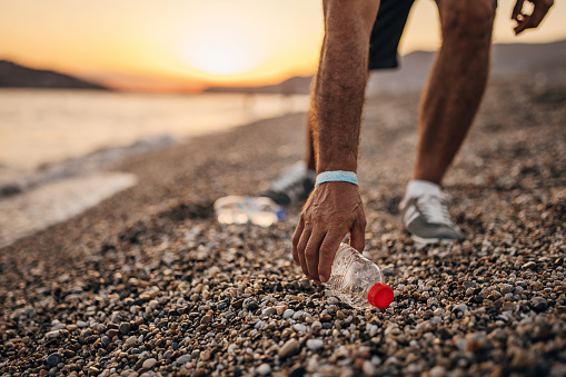 One man, senior male picking up plastic bottle on the beach in sunset. He is cleaning the environment.