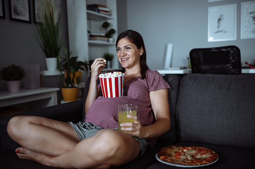 Pregnant woman eating popcorn, pizza and drinking lemonade while watching series on TV