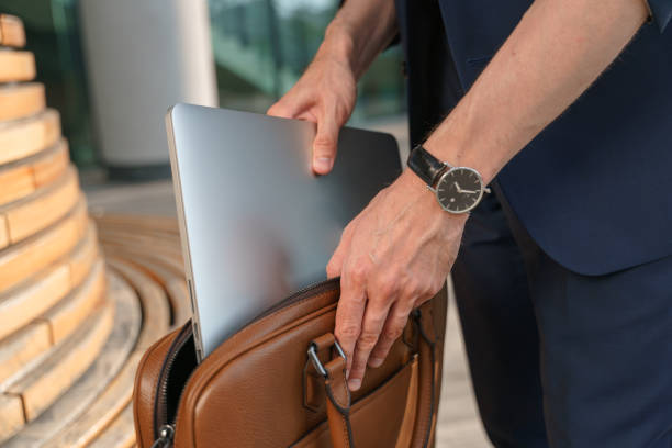 um close-up de um empresário em um terno azul com um bom relógio de pulso está parado perto da bech de madeira contra a fachada moderna de um centro de negócios coloca seu laptop em uma bolsa de couro laptop - bech - fotografias e filmes do acervo