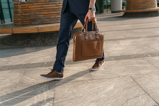 A young man's legs in a blue business suit  in the evening sunlight. The man has a brown leather shoes and a leather laptop bag