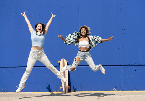 two multiethnic women jumping on a blue wall, with skateboard and casual clothes