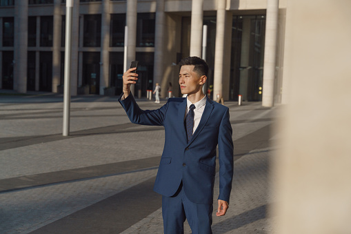 A young asian manager in  a blue suit makes selfie, standing on a square in front of the business center, we see him from the distance in the evening light